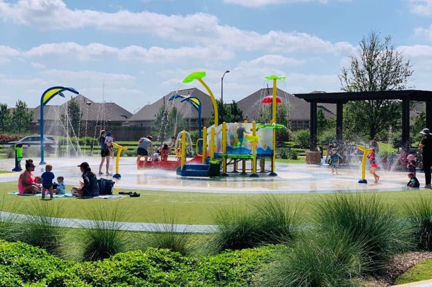 Children playing at the splash pad at Elyson in Katy, TX, with colorful spray features, fountains, and families relaxing on the lawn nearby.