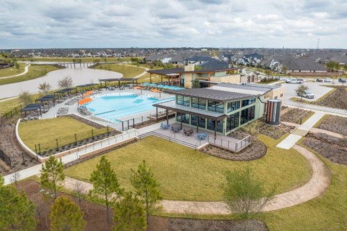 An aerial view of a modern residential community clubhouse with a swimming pool, outdoor seating areas, walking paths, and surrounding suburban homes.